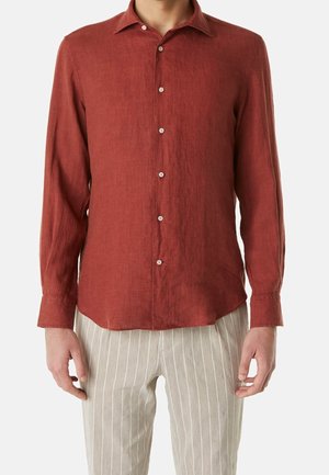 Homme portant une chemise à manches longues couleur rouille boutonnée et un pantalon beige clair à rayures verticales blanches, debout devant un fond neutre.