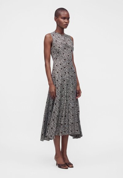 Femme debout portant une robe midi sans manches noire et blanche à motifs floraux et des talons ouverts foncés sur un fond clair uni.
