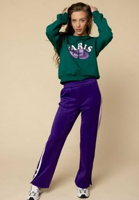 Jonge vrouw in groen "Paris 90" sweatshirt, paarse trainingsbroek met witte strepen, witte sneakers, poserend met handen in haar tegen een beige achtergrond.