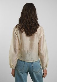 Blusa crema trasparente con maniche lunghe e puffate, con motivi a pois argentati, abbinata a jeans in denim blu a vita alta. Orlo arrotondato.