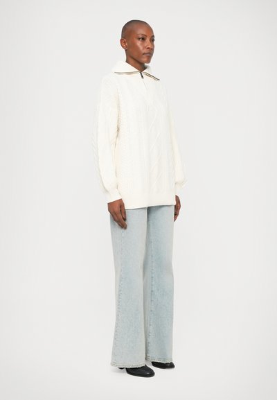 Suéter blanco de punto trenzado con cuello alto, con puños acanalados, combinado con unos jeans acampanados de color azul claro y zapatos negros. Diseño simple y texturizado.