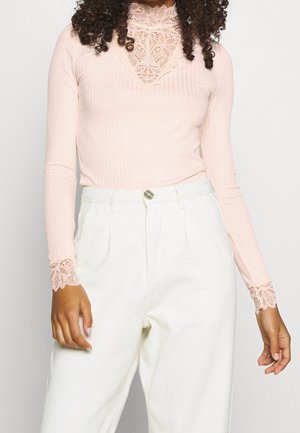 Top a maniche lunghe rosa a costine con colletto in pizzo alto e dettagli in pizzo ai polsini, abbinato a pantaloni bianchi a vita alta.