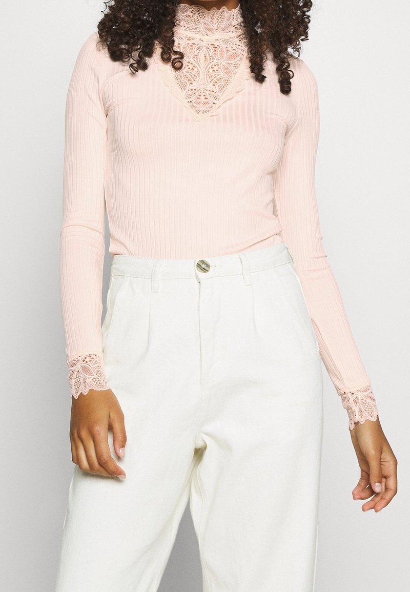 Rosa ribbad långärmad topp med en hög spetskrage och spetsdetaljer vid ärmsluten, ihop med vita högmidjade byxor.