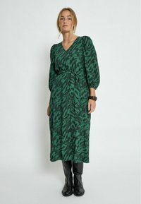 Vihreä kuvioitu mekko, jossa on V-pääntie, puffihihat ja solmittava vyö. Tämä on yhdistetty mustiin nilkkasaappaisiin. Kankaan pinnassa on teksturoitu viimeistely.