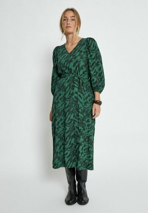 Grünes gemustertes Kleid mit V-Ausschnitt, Puffärmeln und einem Bindegürtel. Getragen mit schwarzen Knöchelstiefeln. Der Stoff hat eine strukturierte Oberfläche.