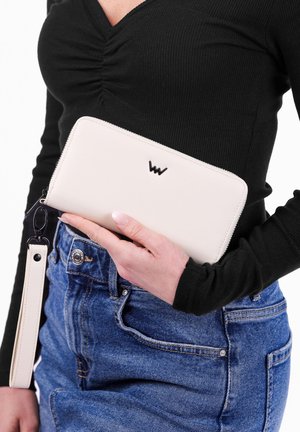 Žena v černém topu a modrých džínách drží bílou kabelku na zip s páskem na zápěstí a černým logem u pasu.