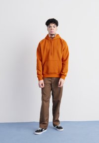 Orange hoodie with a kangaroo pocket and drawstring hood, paired with brown trousers and black sneakers, standing against a light background.