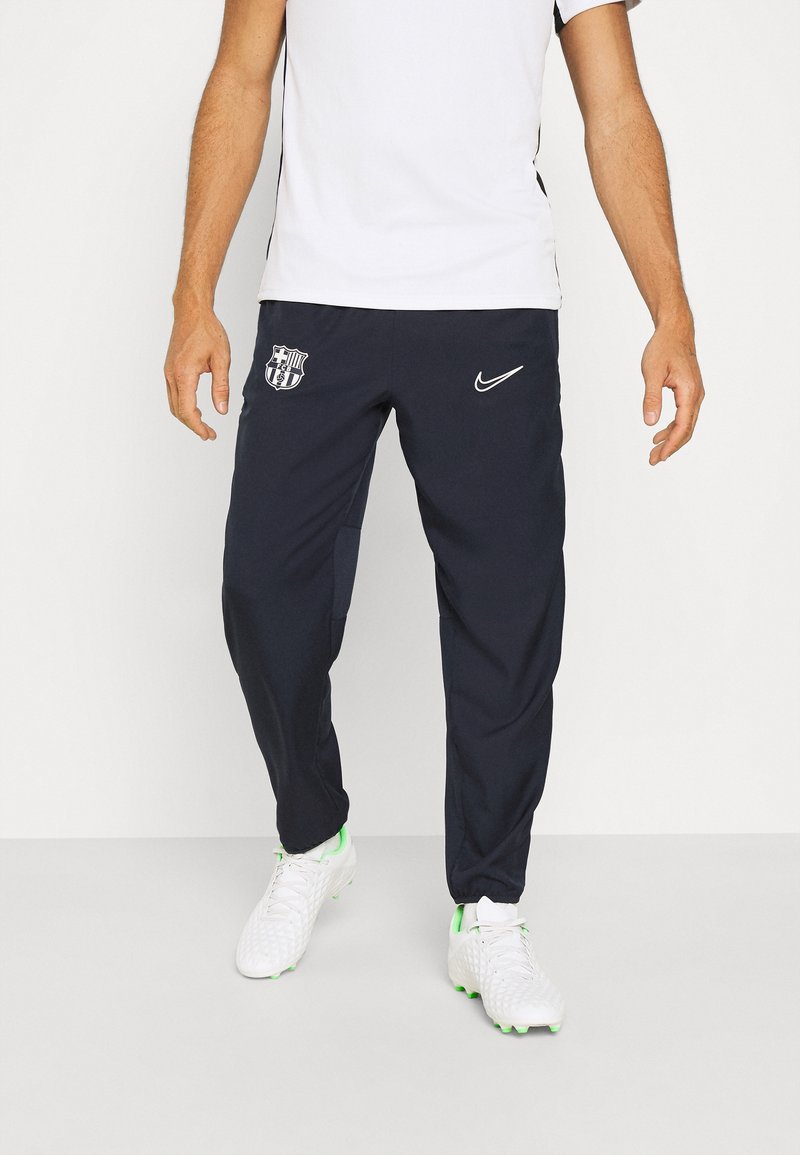 Pantalons de sport navy avec un logo de Barcelona sur la cuisse gauche, dotés d'un design fuselé et de poignets élastiques. Associés à des chaussures blanches.