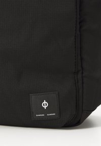 Sort tekstureret taske med et firkantet logo-patch med brandnavnet "SAMSØE SAMSØE" og et cirkulært designelement.