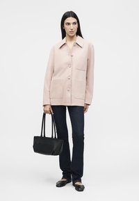 Γυναίκα που φοράει μια ανοιχτό ροζ σακάκι με κουμπιά, σκούρα μπλε τζιν, μαύρες μπαλαρίνες, κρατώντας μια μαύρη τσάντα σε απλό λευκό φόντο.