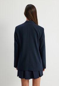 Granatowa marynarka o wyraźnej konstrukcji, z pojedynczymi guzikami na mankietach i rozcięciem z tyłu. Połączona z granatową plisowaną spódnicą, prezentującą monochromatyczny wygląd.