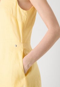 Rochie galbenă din in, evidențiind un design fără mâneci, buzunare laterale și detalii de cusătură subtile, cu o textură netedă și ușoară.