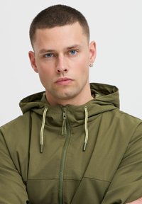 Hanorac verde olive cu fermoar, dotat cu un glugă ajustabilă cu șnur, având o textură netedă și detalii subtile de cusătură.