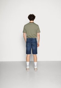 Un uomo dai capelli ricci sta in piedi di fronte a un muro bianco, indossando una maglietta a righe verdi e nere, pantaloni corti di jeans blu, calzini bianchi e sneakers.