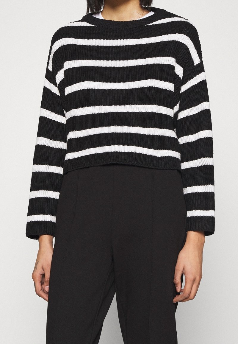 Personne portant un pull à manches longues à rayures horizontales noires et blanches et un pantalon noir taille haute, debout devant un fond uni.