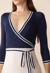 Bodysuit en maille marine avec un décolleté en V profond, des accents rayés blancs et une taille à nouer, associé à une jupe blanche plissée.