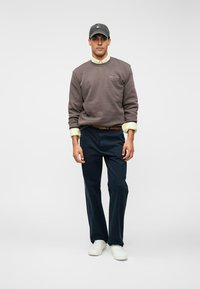 Uomo che indossa una felpa grigia, una camicia beige con le maniche arrotolate, pantaloni blu scuro, sneaker bianche e un cappellino grigio, in piedi su uno sfondo neutro.