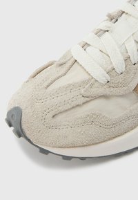 Béžová semišová teniska s textúrovaným zvrškom, zvýrazneným prešitím a bielymi šnúrkami. Má gumovú podrážku so sivým akcentom na špičke.