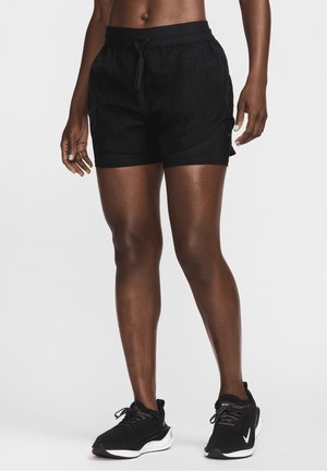 Personne portant un short de sport noir avec cordon et des chaussures de course noires, debout devant un fond blanc uni, vue de la taille vers le bas.