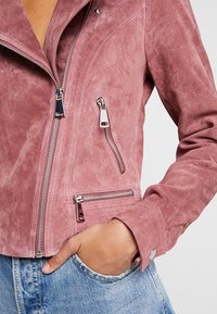 Różowa zamszowa kurtka z srebrnymi zamkami błyskawicznymi i kieszeniami, o dopasowanym kroju i klasycznym kołnierzu. Miękka, teksturowana powierzchnia.