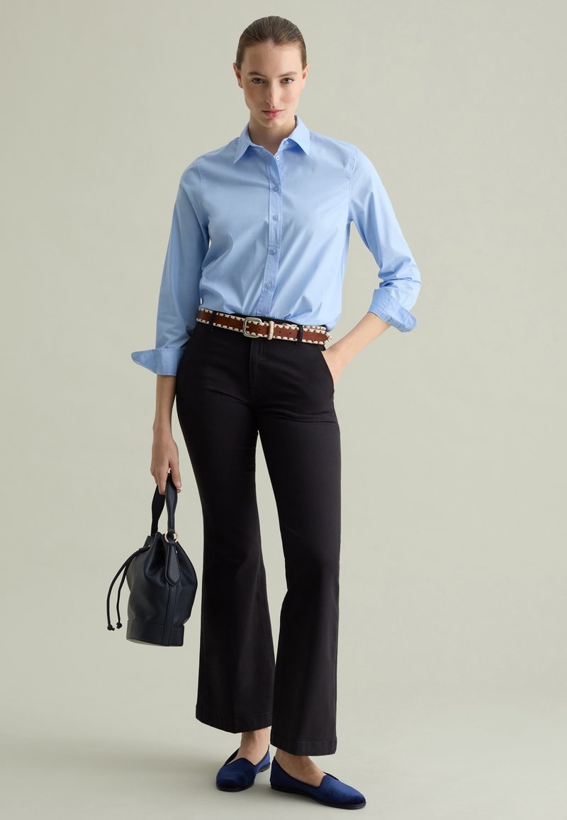 Chemise bleu clair à boutons, pantalon noir évasé, ceinture brune à motifs, ballerines bleu foncé, tenant un sac à dos noir à cordon.