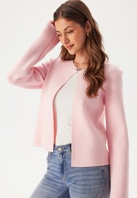 Růžový zkrácený cardigan s dlouhými rukávy, texturovaná látka a zapínání na jeden knoflík. Nositelný přes bílý top a spárovaný s modrými džínami.