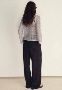 Femme aux cheveux longs et ondulés, debout face au mur, portant un pantalon noir et un pull en maille blanche ajourée, les mains dans les poches.