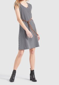 Femme portant une robe rayée verticale noire et blanche avec une ceinture tressée marron et des bottines noires, debout devant un fond uni.