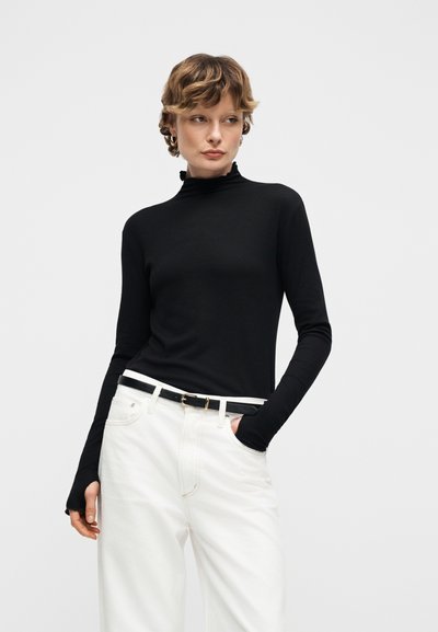 Γυναίκα που φοράει μαύρο μακρυμάνικο πουλόβερ ζιβάγκο και λευκό ψηλόμεσο παντελόνι με μαύρη ζώνη, στέκεται με το ένα χέρι στην τσέπη.