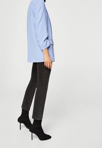 Personne portant un blazer bleu clair, un jean noir slim et des bottines stilettos chaussettes noires, debout devant un fond clair uni.