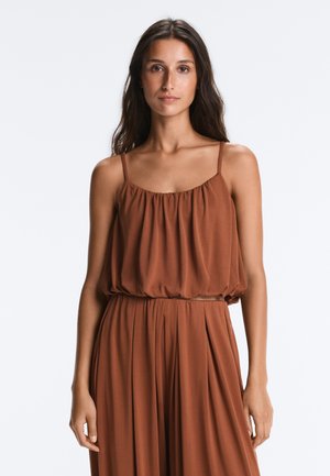 Femme aux longs cheveux foncés portant un haut marron sans manches froncé et un pantalon large taille haute assorti, debout devant un fond uni.