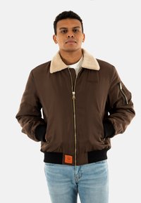 Braune Bomberjacke mit einem krämfarbenen Kunstfellkragen, Reißverschluss, gerippten Bündchen und Saum, seitlichen Taschen und einem orangefarbenen Etikett.