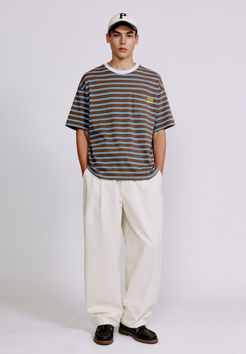 Jeune homme portant un t-shirt rayé marron et bleu, un pantalon blanc large, des chaussures noires, et une casquette de baseball blanche avec une lettre P noire.