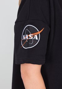 Crna tkanina majica s kratkim rukavima s izvezenim NASA logom na rukavu, s narančastim naglascima i kružnim dizajnerskim elementima.