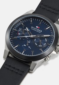 Reloj analógico con un dial texturizado azul, marcadores de hora plateados y tres subdiales. Presenta una correa de cuero negro y una caja de acero inoxidable.