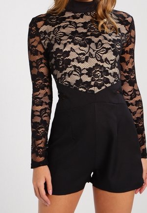 Vrouw die een zwarte kanten top met lange mouwen en bloemmotieven draagt, gecombineerd met zwarte shorts met hoge taille, tegen een effen achtergrond.