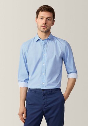 Camisa de botones azul claro con un patrón texturizado, con mangas largas arremangadas en los puños. Combinada con pantalones oscuros de color marino.