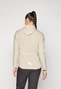 Veste légère beige avec capuche, taille élastique et poignets ajustables ; présente un détail de logo triangulaire à l'arrière.
