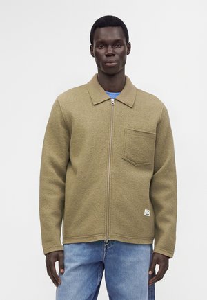 Mand iført beige lynlåstrøje med brystlomme og lyseblå jeans, stående mod en ensfarvet lys baggrund.