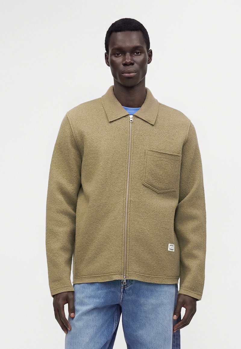 Homme portant une veste beige zippée avec poche poitrine et un jean bleu clair, debout devant un fond uni clair.