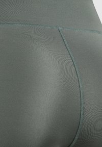 Legginsy z wysokim stanem w stonowanym zielonym odcieniu, wykonane z gładkiego, elastycznego materiału o subtelnym połysku i płaskich szwach dla wygody.