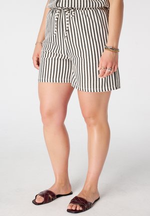 Vrouw draagt zwart-witte korte broek met verticale strepen en kleine stippen, bruine slip-on sandalen en armbanden aan haar rechterpols.