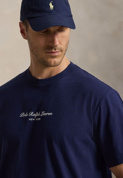 Άνδρας φοράει μπλε σκούρο μπλουζάκι Polo Ralph Lauren και αντίστοιχο καπέλο, κοιτάζοντας προς τα κάτω σε απλό φόντο.