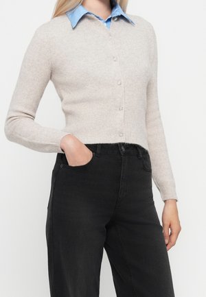 Femme portant un cardigan beige boutonné par-dessus une chemise bleue à col, jean noir taille haute, une main dans la poche.