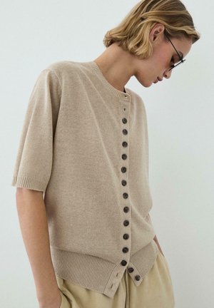 Persona che indossa un cardigan beige a maniche corte con bottoni neri sul davanti, mani nelle tasche dei pantaloni beige, guarda verso il basso, sfondo semplice.