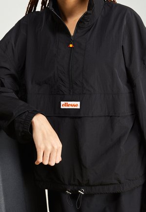 Person wearing black ellesse pullover jacket with half-zip and drawstring hem, arm bent at waist, against neutral background.