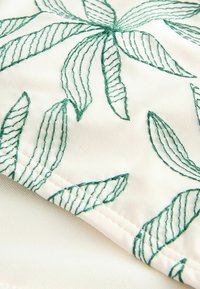 Tissu blanc avec broderie en fil vert formant des motifs de feuilles avec des détails de couture visibles.