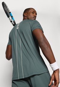 Tume roheline sportlik T-särk lühikeste varrukatega, tekstuuriga kangas, valged aktsentjooned seljal, kantud inimesel, kes hoiab tennisereketit.