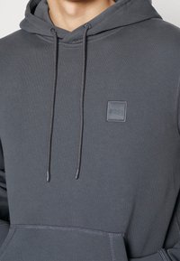 Felpa con cappuccio grigio scuro realizzata in tessuto morbido, dotata di tasca frontale, cordini regolabili e una piccola etichetta con logo sul petto.
