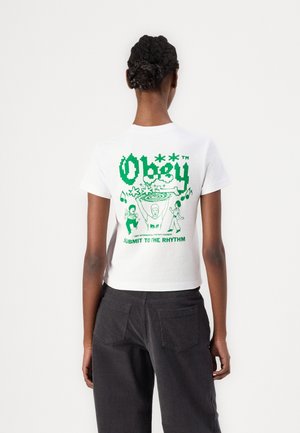 Person wearing white t-shirt with green graphic showing dancing figures, musical notes, and text "Obey" and "Submit to the rhythm" on the back.